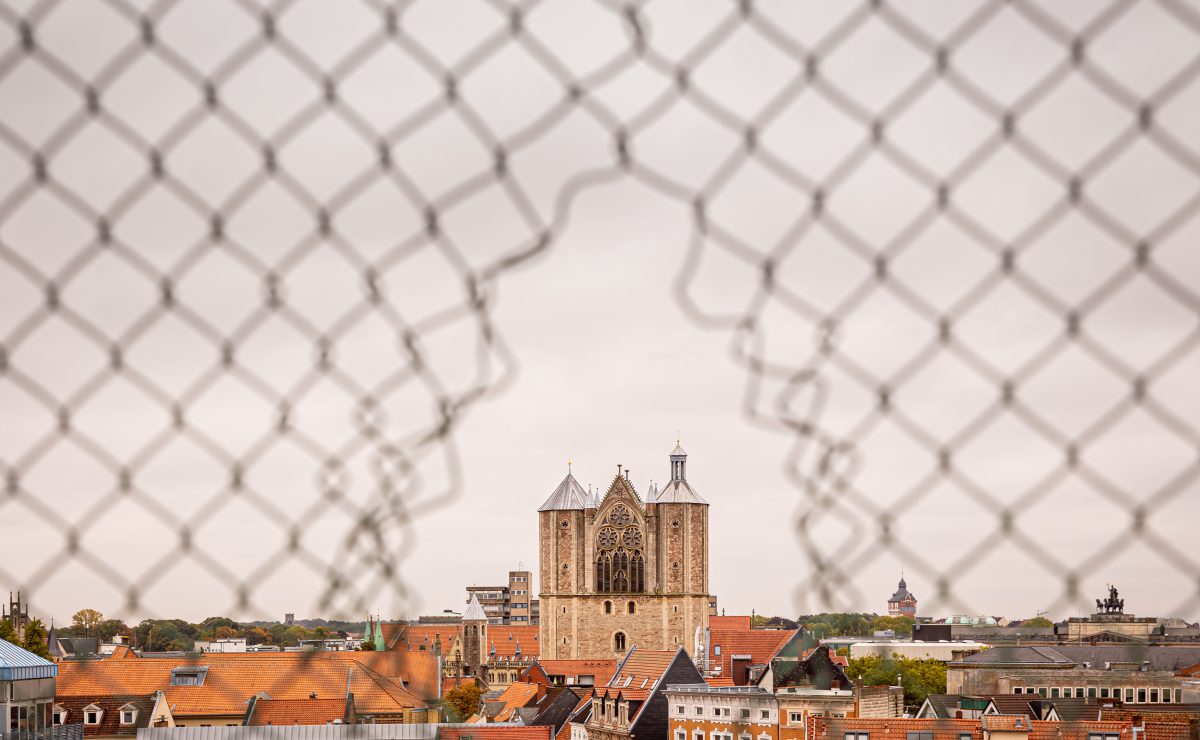 Der Braunschweiger Dom ist bei trübem Wetter durch ein Loch in einem Zaun am Horizont zu sehen.