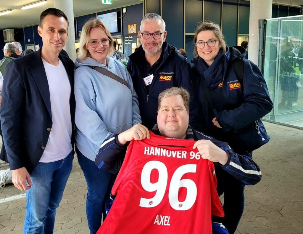 Haben eine dicke Überraschung für Krebspatient Axel im Gepäck: Sportdirektor Marcus Mann und Nadine Große, Assistentin der sportlichen Leitung, überreichen ein personalisiertes Trikot und einen Fanschal. Unsere Wunscherfüller:innen Ralf und Alina freuen sich für ihren schwerkranken Gast.