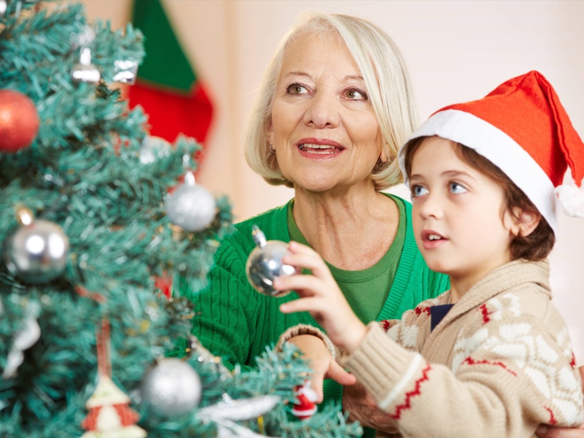 Ein kleiner Junge mit Weihnachtsmann-Mütze schmückt mit seiner Oma einen Weihnachtsbaum.