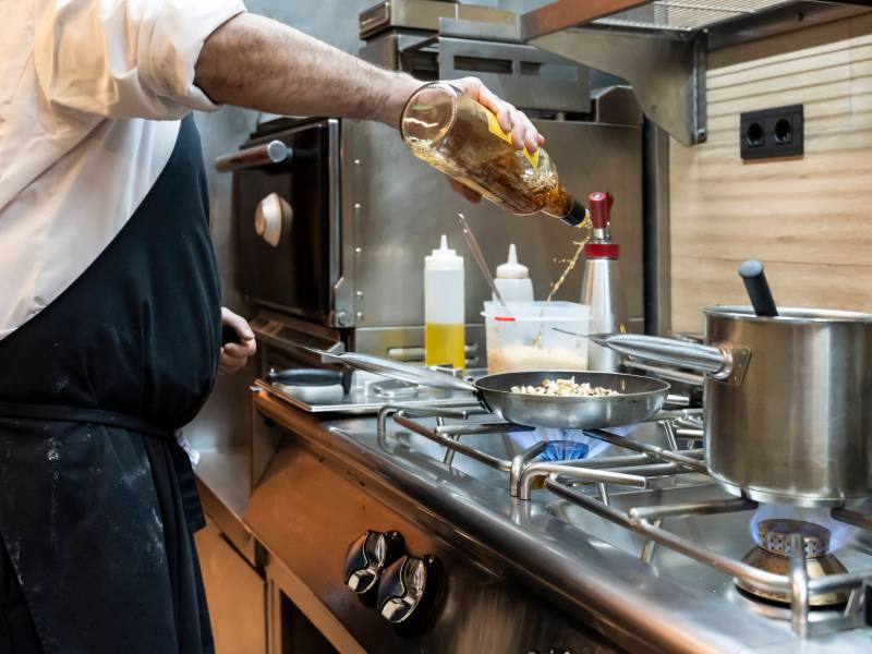 A chef in a professional kitchen is focused on pouring alcohol into a pan,